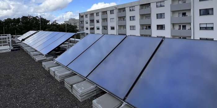 Eine Reihe von Solarpanels ist auf einem Flachdach montiert, mit einem Blick auf ein mehrstöckiges Wohnhaus und grüne Bäume im Hintergrund.