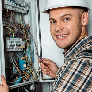 Ein freundlicher Elektriker mit einem Helm lächelt, während er eine elektrische Installation überprüft und Werkzeuge in der Hand hält.