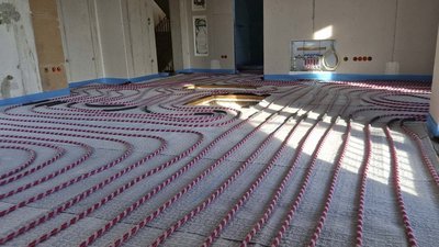 Die Fußbodenheizung ist installiert, mit langen, gewundenen Rohren, die auf einem grauen Untergrund in einem hellen Raum verlaufen.