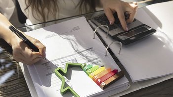 Das Bild zeigt eine Hand, die auf einem Dokument schreibt, umgeben von einem Taschenrechner und bunten Diagrammen.