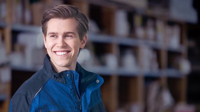 Ein junger Mann mit einem breiten Lächeln trägt eine blaue Arbeitsjacke und steht in einem Lager mit Regalen im Hintergrund.